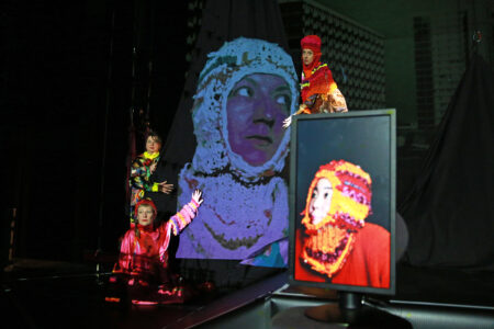 Five performers are on stage, three of whom are physically on stage holding a cloth onto which the image of a performer is projected larger than life. Another person can be seen in the foreground on a computer screen.
