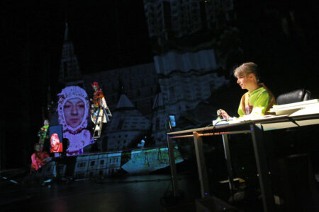 In the mainly dark stage space, the silhouettes of buildings can be seen projected onto the stagewalll. In the foreground, a performer sits at a desk and operates a lever. To the left of the picture is a group of performers.