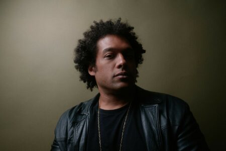 A black man with a prominent chin, short, unkempt afro, black T-shirt and leather jacket in front of a brown-gray background. He looks into the camera from the side with a serious face.