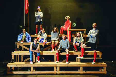 Auf einer hölzernen Tribühne stehen und sitzen neun lächelnde Personen in grauen, roten und blauen Sport-Outfits, mit weißen Schuhen und roten Stulpen an den Beinen.
