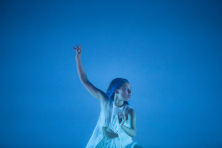 In blauem Licht steht eine Tänzerin in einem weißen, halbtransparenten Kostüm mit Ausschnitt am Bauch und hält einen Arm locker nach oben, der andere Arm vor dem Oberkörper mit angewinkelter Hand. Der Blick geht nach rechts, der Mund steht locker offen.