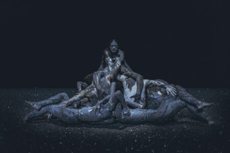 In a dark stage space, completely covered with black glittering rubble, a heap of people piles up. Arms and legs are entangled and everyone is smeared with the black glittering material.