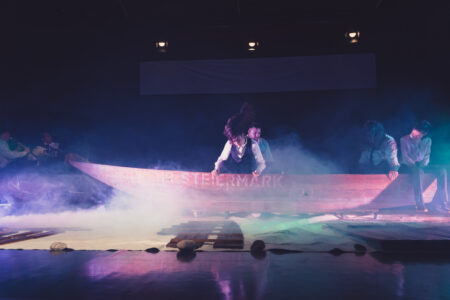 Vier im Hemd gekleidete Personen sitzen auf einem Holzschiff. Zwei in der Mitte und die andere zwei rechts von ihnen. Alle von ihnen schauen auf dem Boden. Die Bühne wird von blauen und pinken Licht bestrahlt. Der Boden der Bühne ist mit Rauch bedeckt.