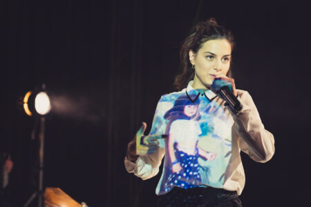 Eine im hellen Hemd gekleidete Frau spricht in ein Mikrofon. Auf dem Hemd der Frau wird ein buntes Bild projiziert.