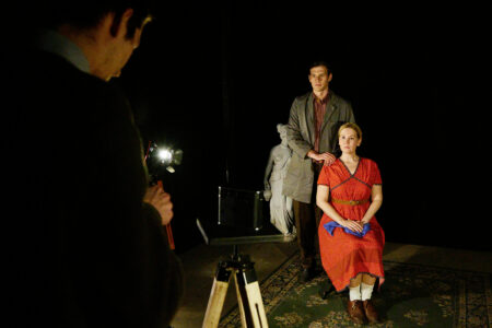 A woman in a red dress is sitting on a chair, behind her is a man in a grey jacket, his hand resting on her shoulder. Both are looking into the camera of a photographer whose back can be seen in the foreground of the picture.