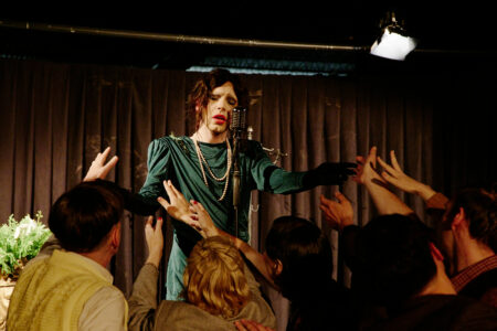 A woman in a green dress in twenties style stands on a stage singing soulfully. With her gloved hands she touches the hands of the people in the audience who reach out to her.