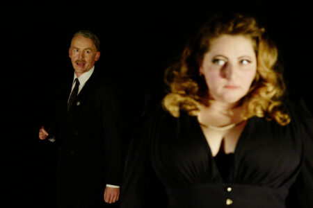 Verschwommenes Foto einer blassen Frau mit blonden, welligen Haaren in schwarzer Kleidung, die skeptisch nach vorne blickt. Im Hintergrund ein Mann in Anzug und Krawatte, der ihr hinterher blickt.