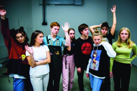 Acht Teenager*innen in einem kargen Raum stehen frontal vor der Kamera in legerer Körperhaltung. Einige von ihnen erheben ihren rechten Arm in die Luft.