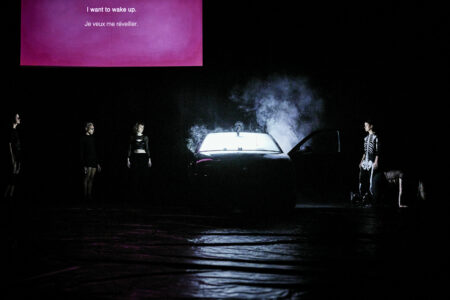 Fünf Personen stehen in einem sehr dunklen Bühnenraum um ein schwarzes Auto herum, aus dem weißer Rauch austritt. Im Hintergrund hängt eine pinke Leinwand mit den Worten "I WANT TO WAKE UP".