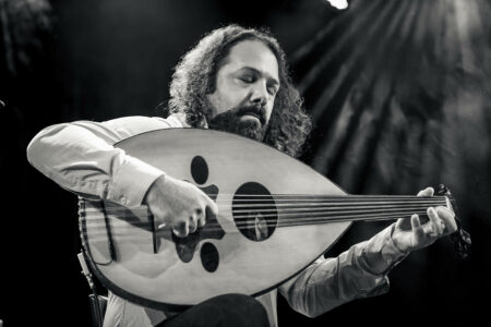 Black and white photo of the musician Wassim Mukdad, a man with a dark beard and curly, shoulder-length hair. He is playing an oud, an Arabic lute, his eyes are closed.
