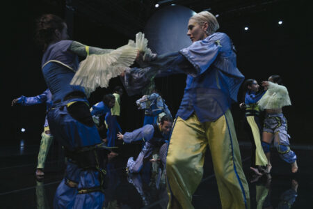 Viele Personen in blauer und gelber Kleidung bewegen sich auf einer dunklen Bühne. Sie tanzen Figuren zu zweit oder allein. Sie stehen teils nah beieinander und halten sich an den Händen.