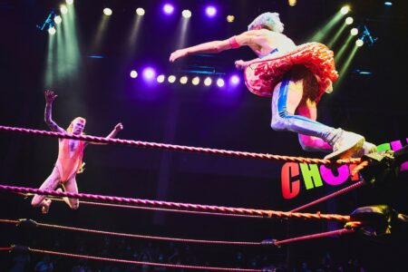 Two drag queens in a fighting ring can be seen. They jump at each other from the corners of the ring. On the wall is written chokehole.