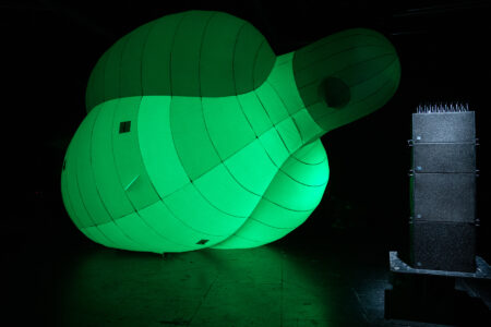 A huge green inflatable pigeon lies tilted to one side on a shiny black floor. Next to it is a tower amplifier with pigeon defence spikes on top.