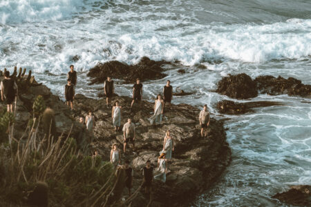 15 Menschen stehen auf einem Fels in der Brandung. Die Frauen und Männer tragen schwarze, weiße und graue Gewänder. Sie haben die Arme an ihrem Körper anliegend. Das Meer schäumt sich auf zu Gischt. Im Wasser sieht man Seetang und in vorne links das Land.