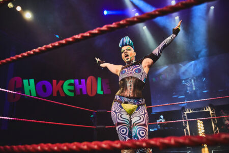 A person with a blue mohawk hairstyle is standing in a fighting ring. She has both hands in the air and looks like she is screaming. In the background on the wall is written Chokehole.