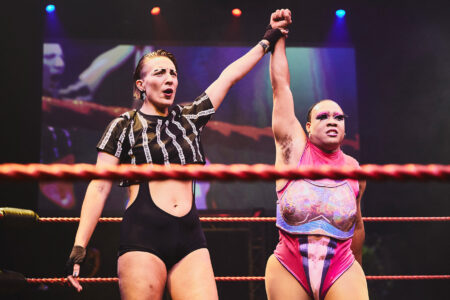 Asset You see two drag queens in a fighting ring. The left one is a referee, the right one is a fighter. The left person holds up the arm of the right, as if she is declared the winner.