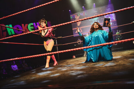 Two drag queens can be seen in a fighting ring. The queen on the left has a black and pink costume and grimaces. The queen on the right has a long blue robe on, kneels on the floor and holds a kind of bible in her hand.