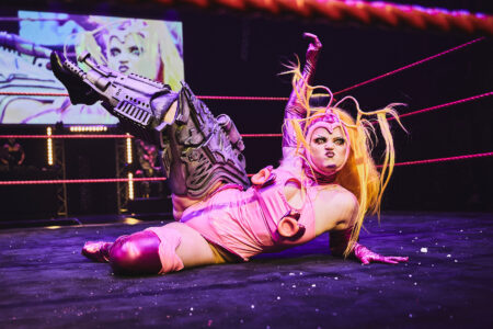 A person is lying in a fighting ring looking at the camera and grimacing. She has a pink costume on and a metal dummy on one leg. Her leg looks like it's from a robot. She stretches the leg upwards.