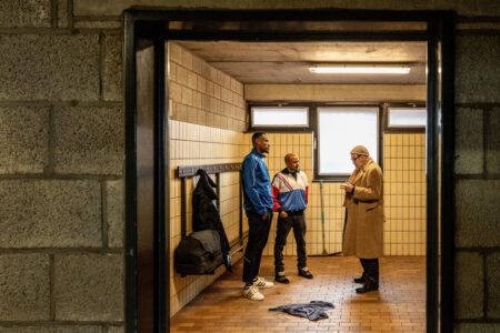 Ein älterer weißer Mann mit beigem Wollmantel und zwei jüngere Schwarze Männer in sportlicher Kleidung stehen in einer gefliesten Umkleidekabine. An einem Haken an der Wand hängt eine Sporttasche, auf dem Boden ein Handtuch.