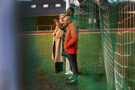 Ein älterer weißer Mann und zwei jüngere Schwarze Männer in Winterjacken und Schals stehen in einem Fußballtor auf einem hellgrünen Rasen. Sie lachen miteinander, einer klatscht in die Hände.