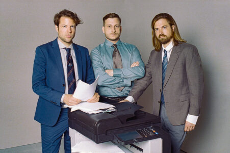 The three members of Brandt Brauer Frick are wearing suits and ties, standing around a copier and looking sternly into the camera. The man on the left has a stack of sheets in his hand, the one in the middle has his arms folded in front of his chest.