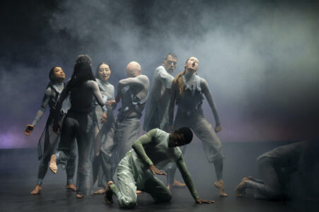 8 people in blue-grey, futuristic costumes dance together in the fog. Their faces are partially distorted, they are grouped close together. One person kneels on the floor, another crawls out from the edge of the picture.