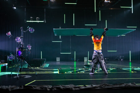 A futuristically dressed person stands wide-legged with hands raised in a dark stage space in which green neon tubes hang vertically and horizontally. Lines are stuck on the floor with green tape, and on the left is a construction of glass spheres.