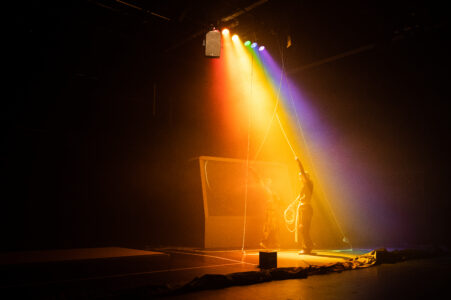 In einem dunklen Bühnenraum stehen zwei Personen, die von regenbogenfarbenen Scheinwerfern beleuchtet werden. Sie stehen vor einer großen Box und ziehen an Seilen, die an den Scheinwerfen hängen.