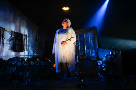 Vor einem in dunkelblaues Licht getauchten Bühnenbild eines Raumes steht in weißem Nachthemd eine dicke Schwarze Frau. Direkt über ihrem Kopf ist ein kleiner Lampenschirm, mit erhobenem Kopf und ihrer linken Hand angewinkelt, spricht sie in den Raum.