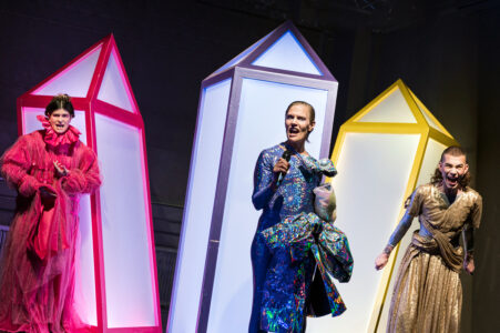 Drei Personen in bunt-glitzernden Kostümen stehen vor diamantenförmigen, spitzen Bühnenelementen in Neonfarben. Die Personen blicken ausdrucksstark zum Publikum. Die Person in der Mitte hält ein Mikrofon in der Hand.