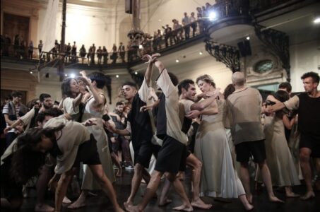 Eine große Gruppe von Menschen tanzt in einem Saal.  Die Tänzer*innen nehmen alle verschiedene Posen ein. Der Raum hat eine Galerie auf der Zuschauer*innen stehen.
