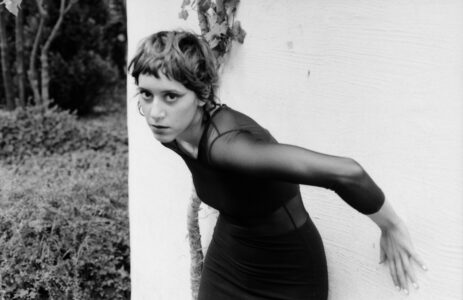 Black and white photo of young slim woman with short hair and nose piercing. She wears a black dress with transparent sleeves and supports herself with her hands on a white wall. Next to the wall are bushes and trees.