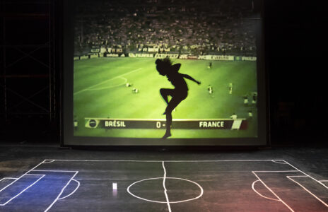 Auf einem dunklen Bühnenboden ist ein Fußballfeld mit kreide eingezeichnet. Im Hintergrund wird ein Fußballspiel auf eine Leinwand projiziert, der Rasen leuchtet grell grün. Vor der Leinwand springt eine Person mit angewinkeltem Bein hoch in die Luft.