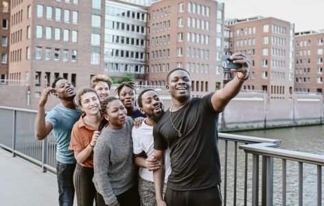 Seven people of different colours take a group selfie with a smile. They are standing on a bridge in front of a modern grey row of houses.