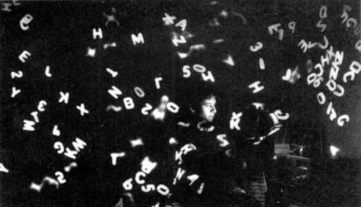 A very dark, grainy black and white photo in which an overexposed face is surrounded by white letters flying all around the room.