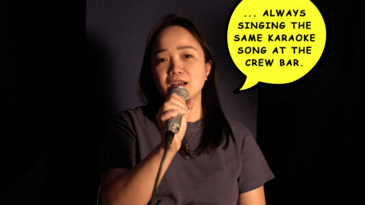 Eine philippinische Frau mit glatten und grauem Tshirt schwarzen Haaren spricht in ein Mikrofon. Neben ihr schwebt eine gelbe Sprechblase in der steht "...always singing the same karaoke song at the crew bar".
