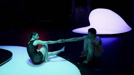 Zwei Personen sitzen in einem dunklen Raum mi lila und türkis beleuchteten rundlich-geschwungenen Bühnenelementen sitzen zwei Personen, die sich distanziert eine Hand reichen und sich intensiv anblicken.
