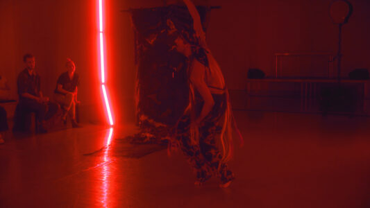In a dark room, lit only by a row of red neon tubes, a man with patterned trousers and a durag in matching fabric moves about. In the background is a large bulging object with two spectators sitting on a bench next to it.