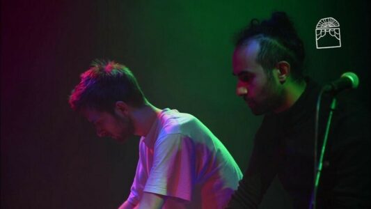 Two young men with beards stand leaning forward with a microphone stand in a very dark room with green and purple light. The one on the left wears a white T-shirt and has short hair, the one on the right has a small bun and wears a black jumper.