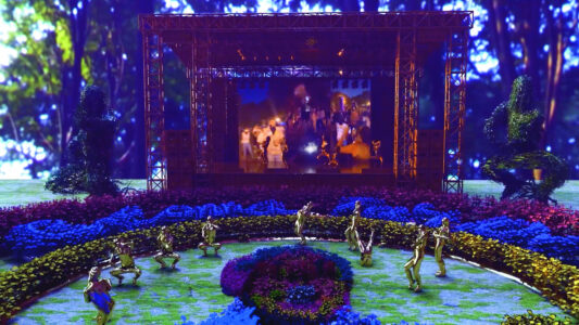 Screenshot eines Videos von einer virtuellen Welt, in der vor einer Leinwand runde Hecken und Büsche in Lila, Blau und Grün wachsen. In einem Hecken-Halbkreis tanzen neun Personen in gold-glänzenden Ganzkörperanzügen.