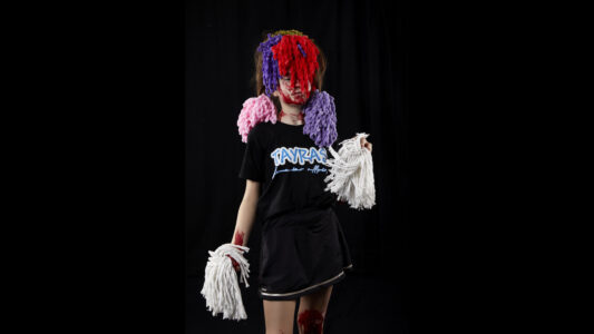 A woman in a black cheerleader costume with Tayras written on it. Two pom-poms in her hands, on her shoulders and in front of her face. Her body is covered in blood.