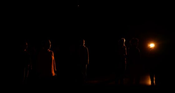 Sechs Menschen werden durch einen Scheinwerfer angestrahlt, sodass nur ihre Silhouetten erkennbar sind. Sie stehen verteilt auf einer Bühne. Sehr dunkler Hintergrund