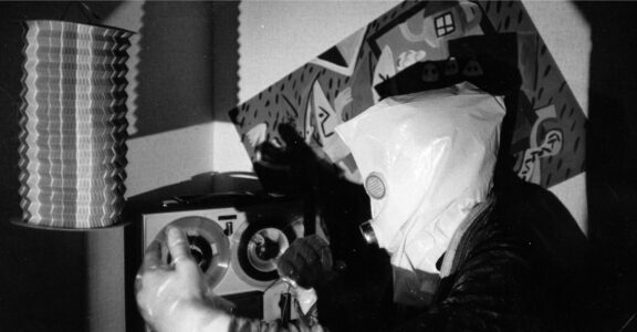 A grainy, slightly blurry black and white photo of a person carrying a white bag over their head. In place of the mouth and eyes are metal discs. The person is reaching for an old tape recorder. In the background hangs an abstract picture.