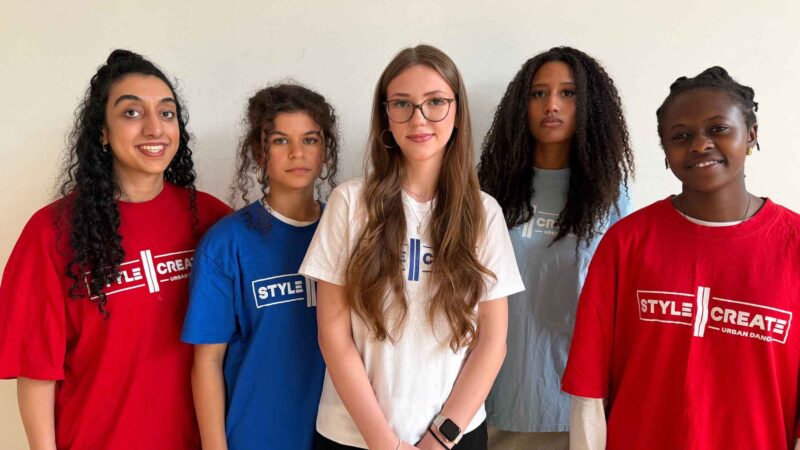 Five people with different hair lengths and textures are wearing colorful T-shirts with the words “STYLE CREATE” printed on them and are standing in front of a light-colored wall.
