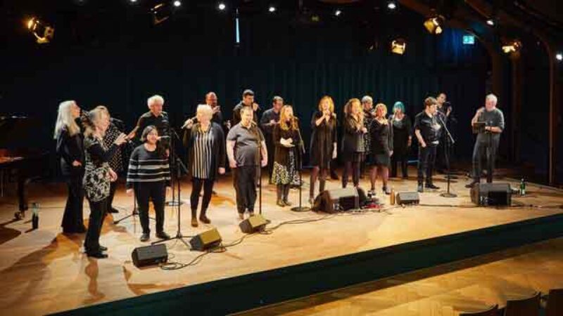 Eine Gruppe von Menschen steht auf einer Bühne und singt gemeinsam in Mikrofone.