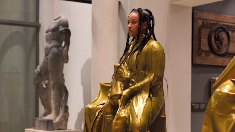 Eine Person in einen glänzenden goldfarbenen Anzug sitzt als Ausstellungsstück im Museum.
