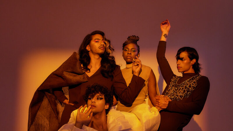 Four People of Colour are standing in elegant poses in front of a brown background with warm yellow lighting. They wear voluminous costumes with balloon sleeves, long skirts and have glamorously styled hair.