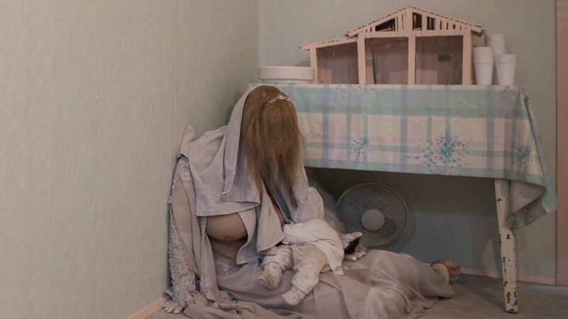 Eine Person mit langen Haaren sitzt mit einem Baby auf dem Boden neben einem Tisch mit einem Modellhaus.