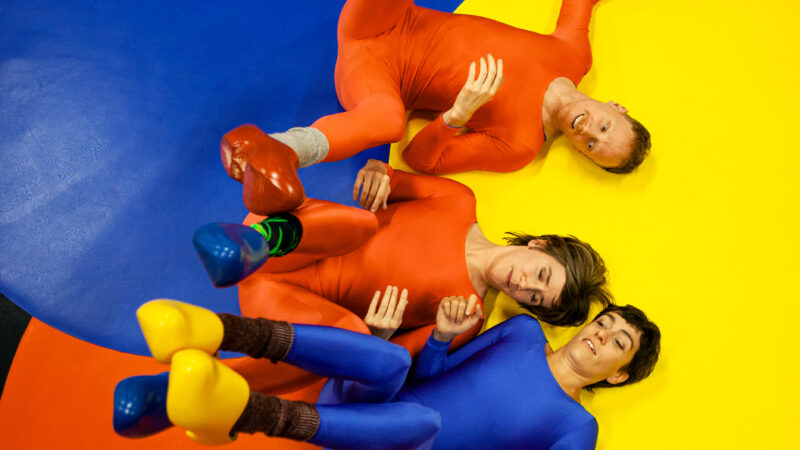 Drei Personen in eng anliegenden, glänzenden Ganzkörperanzügen in rot und blau liegen auf rot-blau-gelben Kreisen auf dem Boden und strecken Arme und Beine in die Luft.